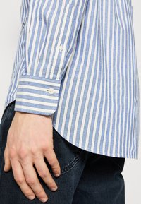 Main masculine reposant sur le côté, portant une chemise à manches longues rayée verticale bleue et blanche avec des boutons aux poignets et un jean bleu foncé.