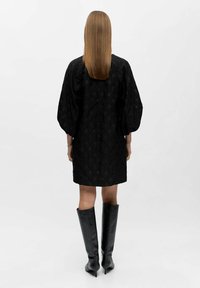 Kvinde med glat brunt hår iført en sort tekstureret kjole med ballonsleeves og sorte knæhøje hæle støvler, stående mod en hvid baggrund.