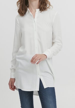 Vrouw die een lange witte blouse met knopen en kraag draagt en een donkerblauwe jeans, staand tegen een effen lichte achtergrond.