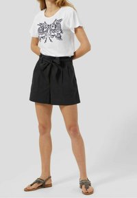 T-shirt blanc avec un design brodé noir, associé à un short noir taille haute avec ceinture à nouer ; sandales marron avec des accents décoratifs.