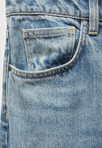 Denim tkanina v svetlo modri barvi z subtilnim vzorcem bledenja. Ima sprednjo žep, svetlo rjave šive in kovinske zakovice za dodatne podrobnosti.