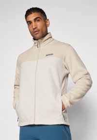 Beige fleecejakke med høj krave, frontlynlås og logoaccent. Har to sidelommer og ribbede manchetter. Glat tekstur.