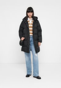 Vrouw in een zwarte pufferjack over een gestreepte trui en een wit shirt, lichtblauwe ripped jeans, zwarte schoenen en die een kleine zwarte handtas vasthoudt.