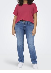 Woman wearing a red short-sleeve top, blue jeans with a black belt, and white sneakers, standing against a plain white background.