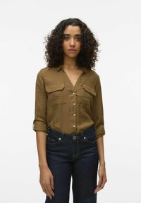 Camisa marrón de botones con mangas enrolladas, dos bolsillos frontales y un diseño de cuello en V. Lleva puesto un pantalón vaquero azul oscuro. Textura de tela suave.