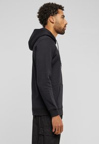 Zwarte hoodie met een kangoeroezak, verstelbare trekkoord en geribde manchetten, weergegeven vanuit een zijprofiel tegen een neutrale achtergrond.