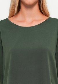Top verde con cuello redondo, mangas largas y tejido liso y texturizado. El diseño es simple y relajado, extendiéndose hasta los hombros.