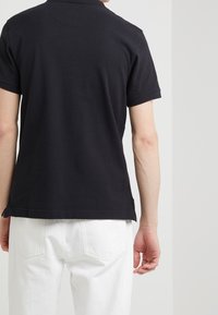 Černé polo tričko s texturovanou tkaninou, krátkými rukávy a klasickým límečkem; nošeno s bílými džíny, což vytváří čistý a minimalistický design.