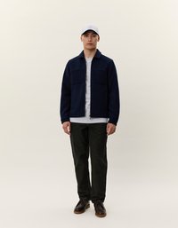 Man standing wearing a white cap, navy zip-up jacket over a white shirt, black pants, and brown shoes against a plain light background.