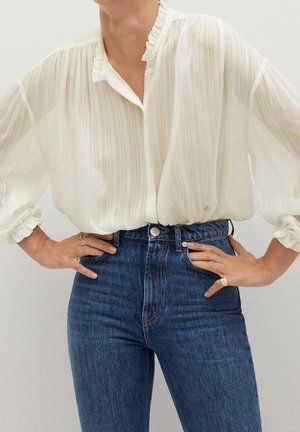 Hvid, let plisseret bluse med rynket krave, kombineret med højtaljede blå jeans. Stoffet virker gennemsigtigt og flydende.