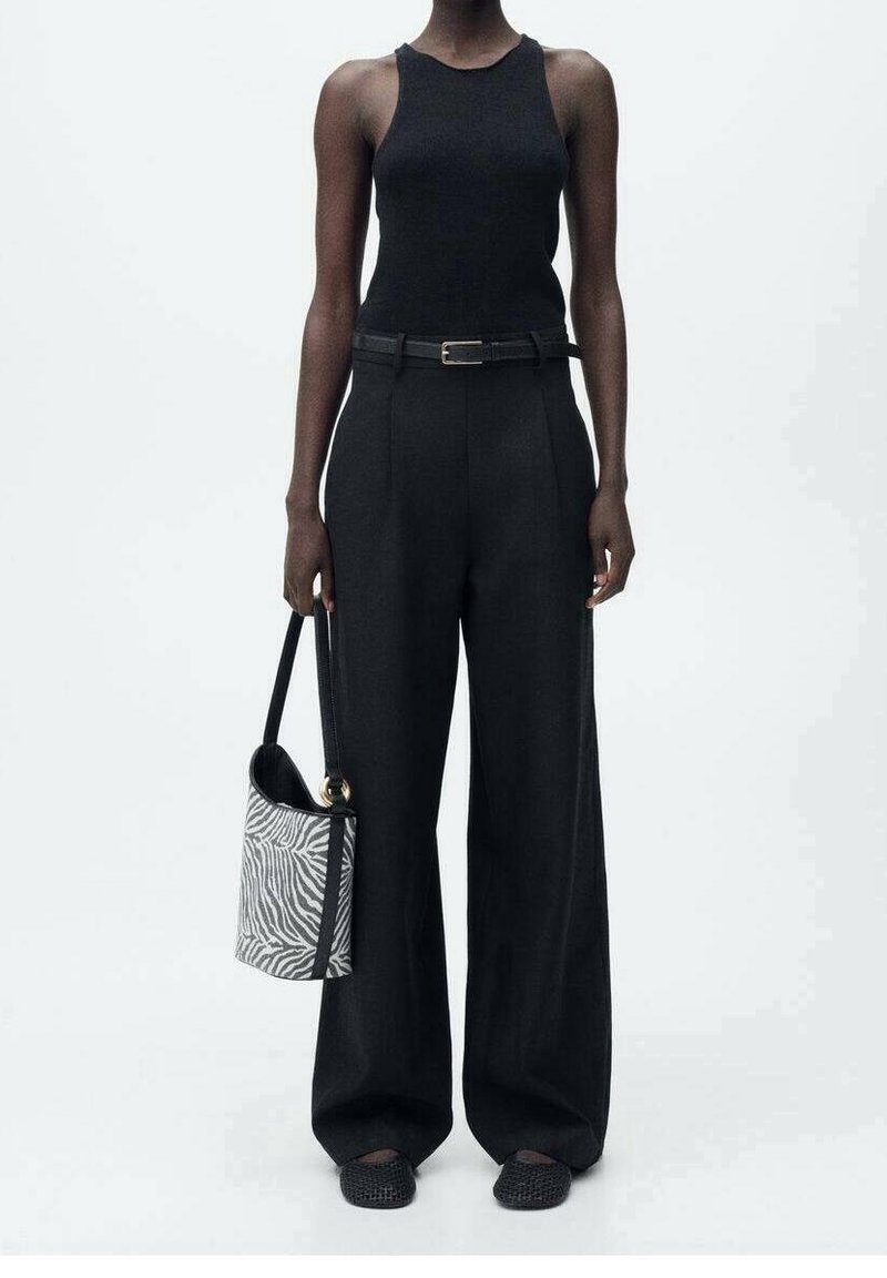 Personne portant un haut noir sans manches et un pantalon à jambes larges tenant un sac à main aux rayures zébrées, debout sur fond blanc.