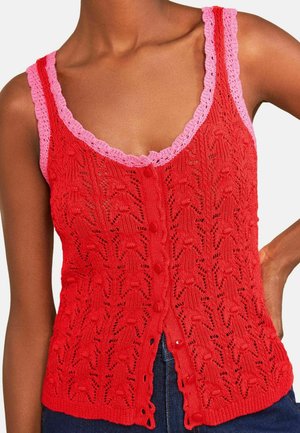 Frau trägt rotes, gestricktes ärmelloses Top mit rosa gezackten Kanten und vorderer Knopfleiste, kombiniert mit blauer Jeans.