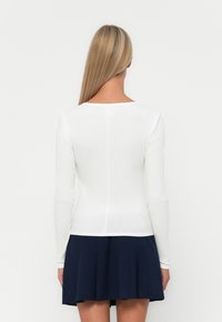 Witte ribstof top met lange mouwen en een ronde halslijn, gedragen met een marineblauwe geplooide rok. De gladde textuur en de aansluitende pasvorm benadrukken de silhouet.
