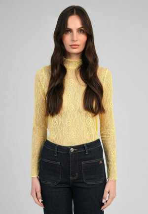 Femme aux longs cheveux foncés portant un haut en dentelle jaune à fleurs à manches longues et un jean taille haute foncé, debout devant un fond gris.