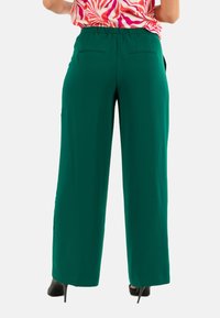 Pantaloni larghi verdi con elastico in vita, texture liscia e due tasche posteriori. Indossati con un top a fantasia colorato.