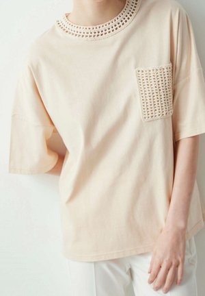 Chemise beige clair à manches courtes avec col en crochet et poche poitrine portée par une personne debout devant un fond uni.