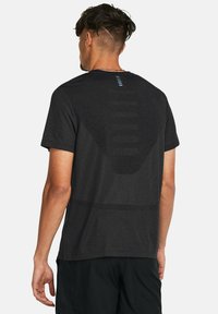 Hombre con cabello corto y oscuro que lleva una camiseta atlética negra texturizada y pantalones cortos negros, de pie de espaldas contra un fondo simple.