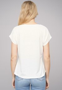 T-shirt blanc à manches courtes avec une coupe décontractée, col rond, texture douce, et un ourlet légèrement arrondi à l'arrière, associé à un jean en denim clair.