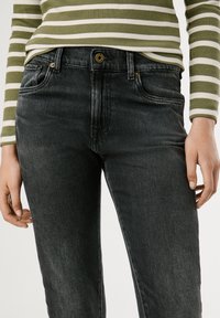 Melni džinsi ar taisnu piegriezumu, kuriem ir zelta piederumi un sānu kabatas, kopā ar zaļu un baltu svītrainu garām piedurknēm kreklu.