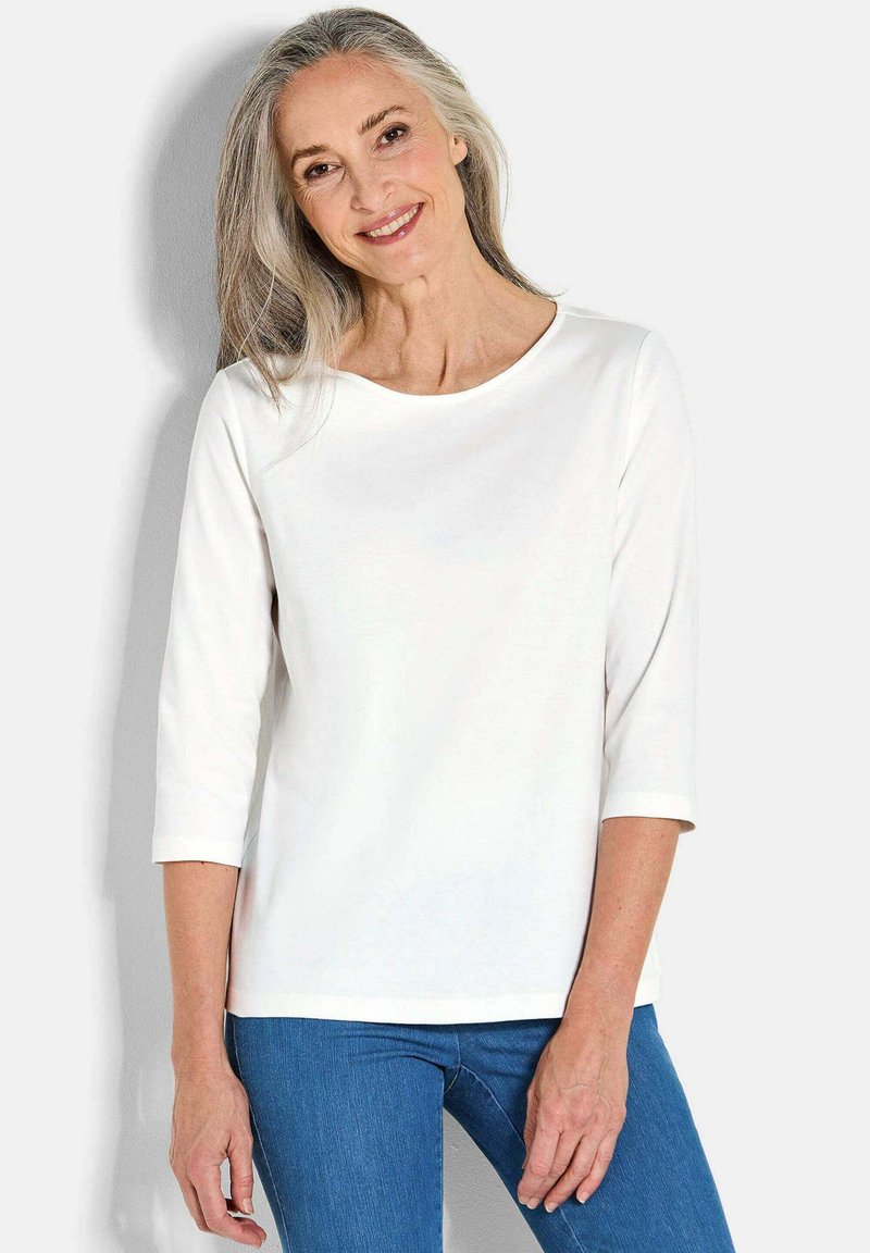 Weiße Langarmbluse mit Rundhalsausschnitt und Dreiviertelärmeln, kombiniert mit blauen Jeans. Glatte Stoffoberfläche, lässiges Design.