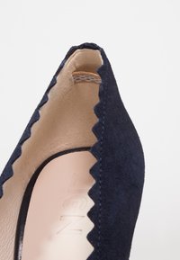 Gros plan d'une chaussure plate en daim bleu marine avec des bords festonnés et une doublure intérieure beige, montrant une partie de la semelle intérieure avec un texte en relief.