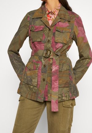 Vrouw draagt een camouflagejack met een ceintuur, bedrukt met roze en olijfgroene bladmotieven, gecombineerd met zandkleurige cargobroek en een overhemd met patroon en kraag.