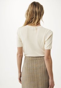 Mujer con largo cabello rubio que lleva una blusa corta de manga en color crema y una falda de cuadros marrón de cintura alta, de espaldas contra un fondo blanco.