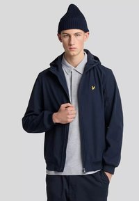 Giacca zip-up blu navy con cappuccio, polsini a coste e logo giallo sul petto; indossata sopra una polo grigia chiara. Il modello indossa anche un berretto blu navy.