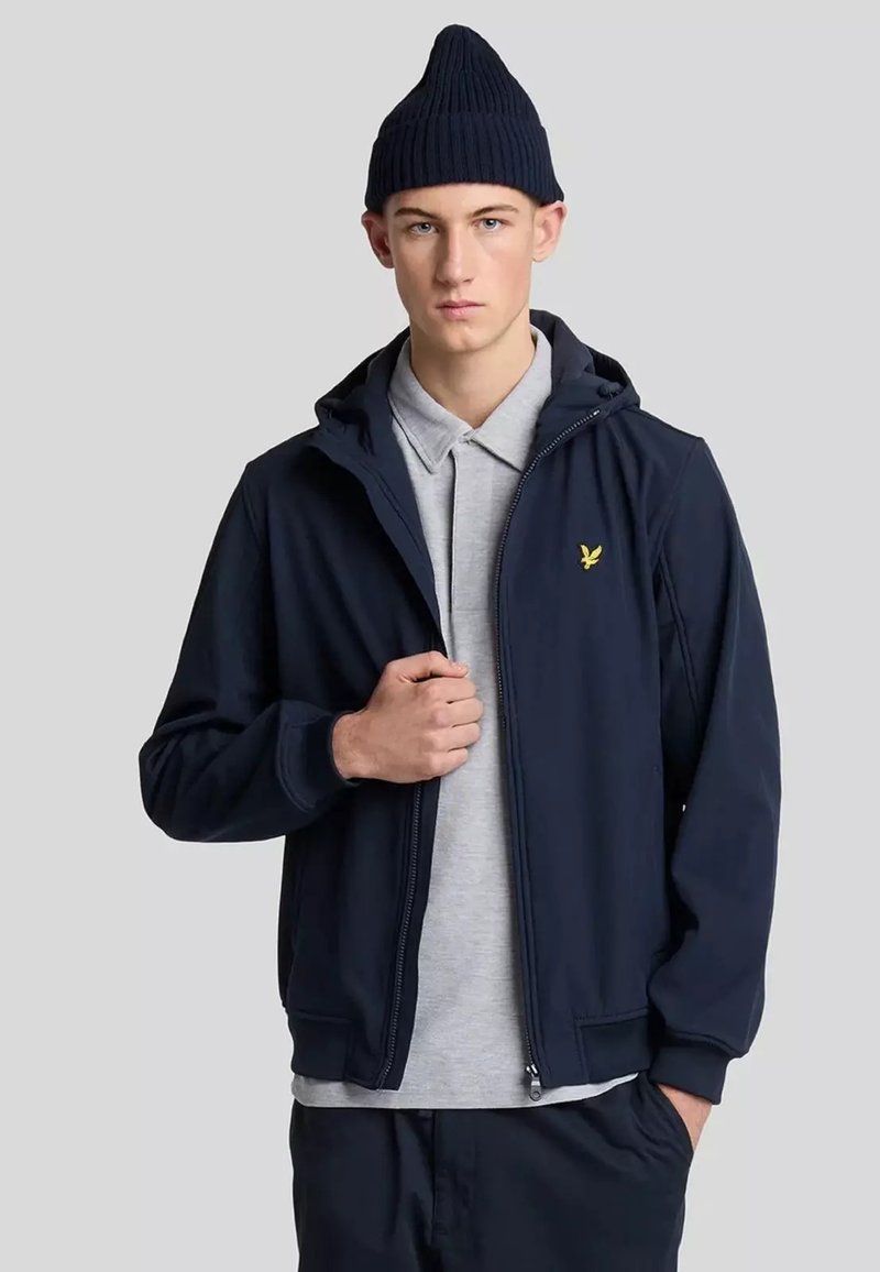 Giacca zip-up blu navy con cappuccio, polsini a coste e logo giallo sul petto; indossata sopra una polo grigia chiara. Il modello indossa anche un berretto blu navy.