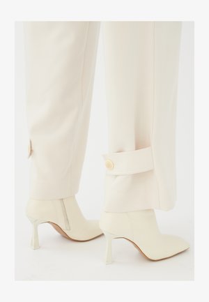 Bež krojene hlače s manžetom, s detaljem gumba, u kombinaciji s bijelim gležnjačama koje imaju prozirni štikli.