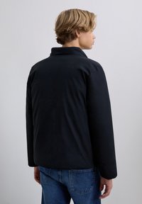 Junge Person mit blonden Haaren, die eine schwarze Jacke und eine blaue Jeans trägt, steht mit dem Rücken zu einer schlichten grauen Hintergrund.