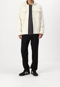 Fleecejacka i krämfärg med två framfickor och svarta tryckknappar, bärs över en mörkstickad tröja och matchas med svarta byxor och sneakers.