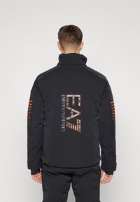 Čierna prešívaná bunda s vysokým golierom, na chrbte s veľkým logom EA7 v metalických farbách a oranžovými pruhmi na rukávoch.