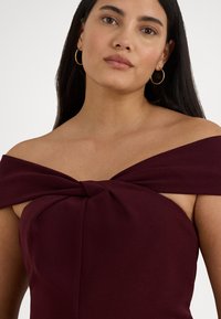 Φόρεμα σε μπορντό χρώμα με σχέδιο χωρίς τιράντες και κόμπο στο μπροστινό μέρος. Κατασκευασμένο από απαλό, υφασμένο ύφασμα με εφαρμοστή σιλουέτα.