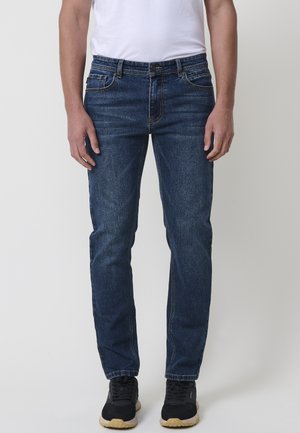 Jeans de mezclilla azul oscuro con un diseño de corte recto, que cuentan con bolsillos frontales y una cremallera, acompañados de zapatillas negras.