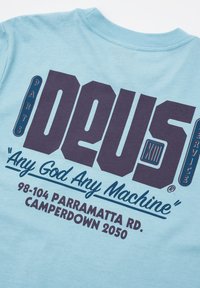 Light blue cotton T-shirt with dark blue and purple graphic text. Features brand name "DEUS" and slogan “Any God Any Machine” in bold fonts.