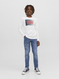 T-shirt branca de manga longa com "JACK & JONES" impresso em vermelho e azul marinho, combinado com jeans ajustados azuis e sapatilhas azul marinho com solas brancas.