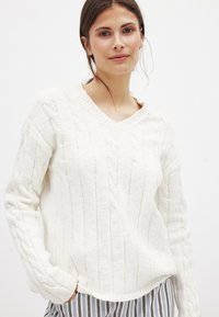 Weißer Strickpullover mit Zopf Muster, V-Ausschnitt, langen Ärmeln und Rippbündchen. Der Stoff hat eine weiche Textur. Das Modell trägt gestreifte Hosen.
