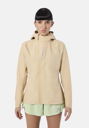 Kobieta o krótkich ciemnych włosach, ubrana w beżową kurtkę z kapturem zapinaną na zamek oraz jasnooliwkowe szorty, stojąca na jednolitym tle.