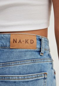 Jeans de mezclilla en azul claro con un parche trasero de cuero texturizado que presenta "NA-KD" grabado en marrón. Incluye costuras en contraste y trabillas para el cinturón.