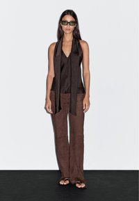 Top in satin marrone con scollo a V e dettagli di foulard annodato, abbinato a pantaloni larghi in suede marrone e sandali bassi su uno sfondo scuro.