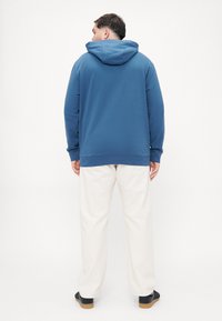 Felpa blu con tasca a marsupio frontale, polsini e orlo a costine. Abbinata a pantaloni a gamba larga color crema e scarpe nere.