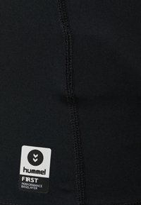 Svart tyg med vertikal söm och en rektangulär svart- och vit etikett som säger "hummel F1RST PERFORMANCE BASELAYER" i nedre vänstra hörnet.