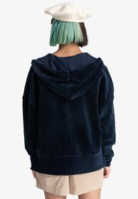 Navy corduroy hoodie with a front kangaroo pocket, large hood, and relaxed fit. Paired with light beige shorts.