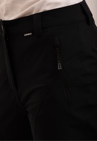 Gros plan sur un pantalon noir avec une poche zippée à l'avant et une petite étiquette de marque au-dessus de la poche, porté avec un haut blanc.