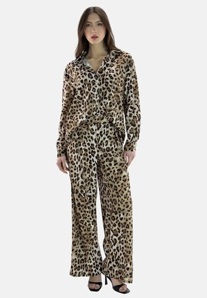 Vrouw die een lange mouwen blouse met luipaardprint en wijd uitlopende broek draagt, gecombineerd met zwarte sandalen met hoge hak, staand tegen een effen achtergrond.