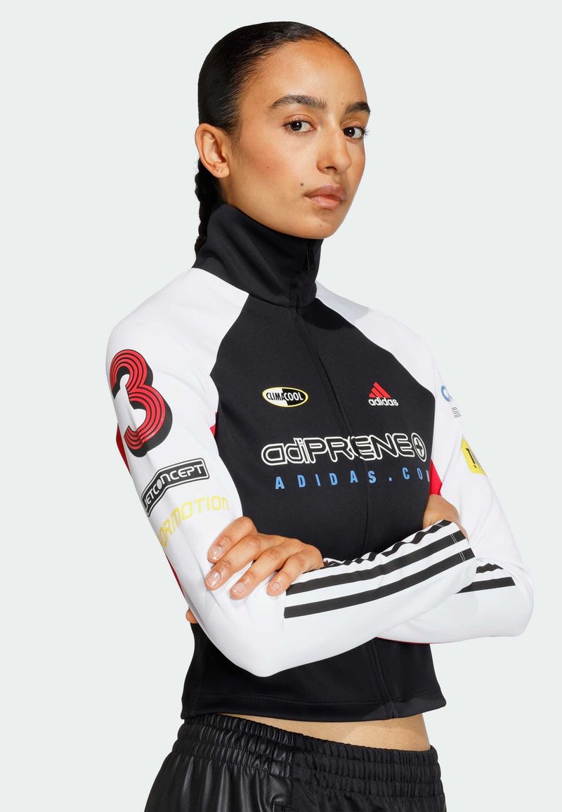 Sort og hvid atletisk jakke med høj krave, røde accenter, flere logoer og stribede ærmer, parret med sorte bukser med elastisk talje.