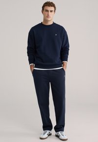 Tamnoplava trenirka majica i pripadajuće hlače izrađene od mekanog tkanog materijala. Majica ima okrugli izrez i mali logotip; hlače imaju bočne džepove. Bijele tenisice.