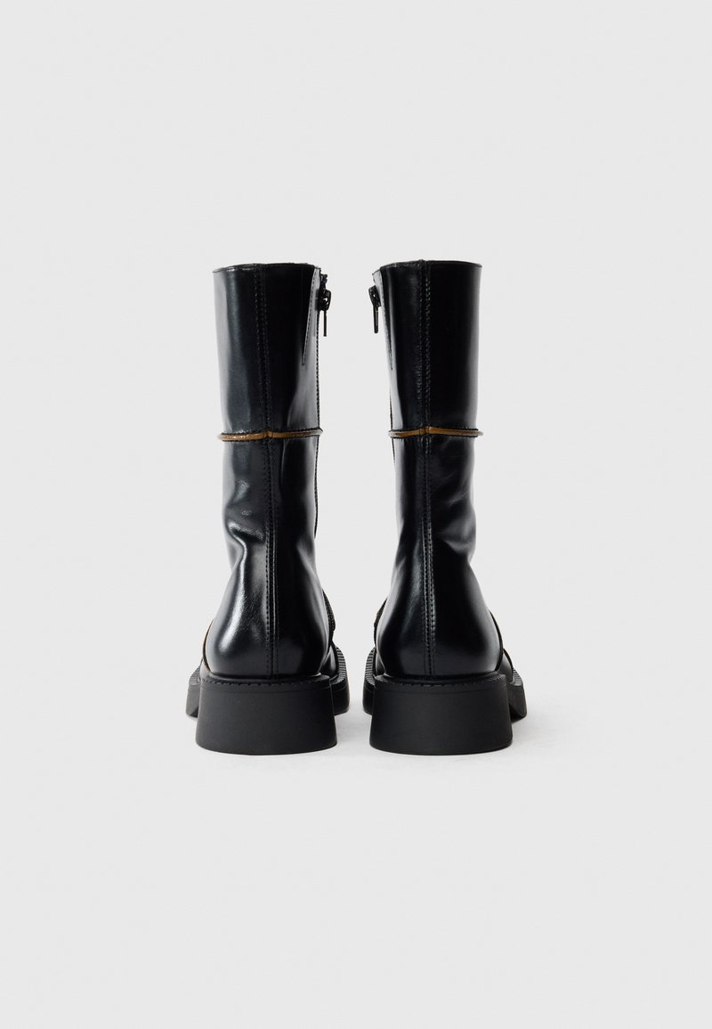 Ett par svarta läderstövlar halvvägs upp på vaden med blixtlås på insidan och tjocka platta sulor, sedda bakifrån mot en vit bakgrund.