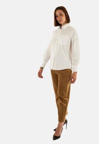 Chemise blanche boutonnée avec une poche poitrine, associée à un pantalon en velours côtelé marron ; le modèle porte des talons hauts noirs, debout sur un fond neutre.